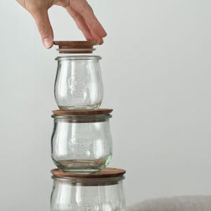 May include: Three clear glass jars with wooden lids stacked. The jars have a rounded shape and a logo on the front. A hand is placing a lid on the top jar. The jars are suitable for food storage.
