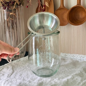 May include: A stainless steel conical strainer with a long handle positioned over a clear glass jar featuring the embossed WECK logo on the front. The strainer has a fine mesh base with a small central post, and the jar has a wide opening.