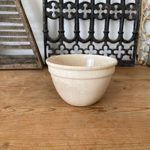 Può includere: Una ciotola in ceramica color crema con una base liscia e arrotondata e un distinto bordo orizzontale vicino alla parte superiore. La ciotola presenta una sottile finitura smaltata a craquelé su tutta la superficie esterna.