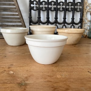 Pode incluir: Um conjunto de quatro taças de cerâmica para pudim em vários tons de branco e creme. Cada taça apresenta uma base lisa e arredondada e uma borda distinta perto do topo, exibindo um design simples e funcional para uso na cozinha.