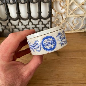 May include: A small, antique ceramic container with a lid, held in a hand. The container is off-white with blue lettering that reads "Beauty" and "Borax". The words "Ho me Sweet Home" are also visible. The container has a vintage aesthetic.