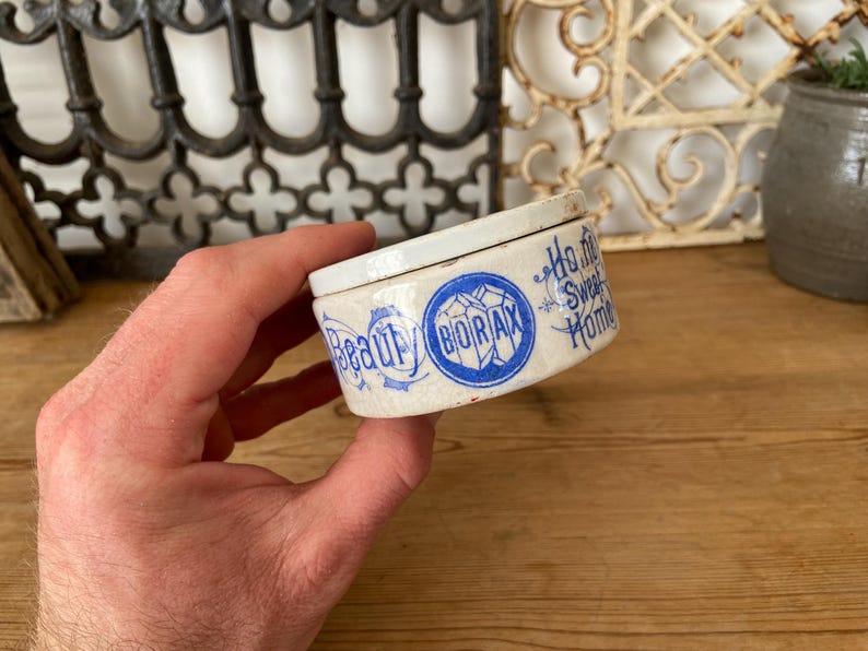 May include: A small, antique ceramic container with a lid, held in a hand. The container is off-white with blue lettering that reads "Beauty" and "Borax". The words "Ho me Sweet Home" are also visible. The container has a vintage aesthetic.