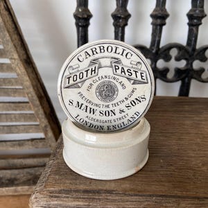 Könnte beinhalten: Ein cremefarbener Keramikbehälter im Vintage-Stil mit einem runden Deckel, der den schwarzen Aufdruck Carbolic Tooth Paste for Cleansing and Preserving the Teeth and Gums, S. Maw Son and Sons, Aldersgate Street, London, England trägt.