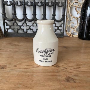 Puede incluir: Un tarro de cerámica de color crema con cuello estrecho y base ancha. El frente presenta un texto impreso en negro que dice Escoffier MOUTARDE AUX FINES HERBES. La superficie tiene un acabado de esmalte craquelado en todo el exterior del tarro.
