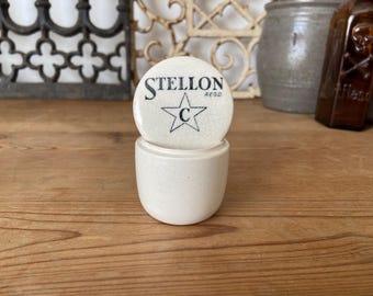 May include: A small, off-white ceramic jar with a matching lid. The lid features the word "STELLON" above a star with the letter "C" inside. The word "REGD" is below "STELLON". The jar is on a wooden surface.