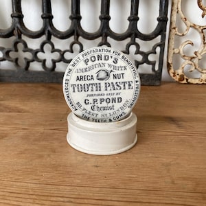 Könnte beinhalten: Ein runder weißer Keramikbehälter mit einem Deckel, der den schwarzen Aufdruck POND'S AMERICAN WHITE ARECA NUT TOOTH PASTE PREPARED ONLY BY G.P. POND CHEMIST 68 FLEET ST LONDON THE BEST PREPARATION FOR BEAUTIFYING & PRESERVING THE TEETH & GUMS trägt.