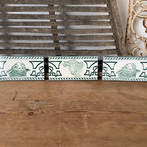 May include: Three vintage ceramic tiles with green and white designs. Each tile features a different animal: a cow, a sheep, and a lamb. The tiles have a decorative border with a repeating pattern.