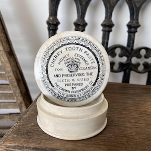 May include: A round, off-white ceramic container for Cherry Tooth Paste. The lid features black text reading: Cherry Tooth Paste, Highly Esteemed, For Cleansing and Preserving the Teeth & Gums, Prepared by The Crown Perfumery Co, 177 New Bond St London.