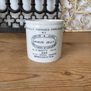Op de afbeelding: Een witte keramische pot met een cilindrische vorm, met een blauw en zwart etiket met de tekst "INVALID JELLY" en "PARKER & SONS". De pot heeft de tekst "18 ST MARYS GATE AND 12 ST ANNS SQUARE MANCHESTER."