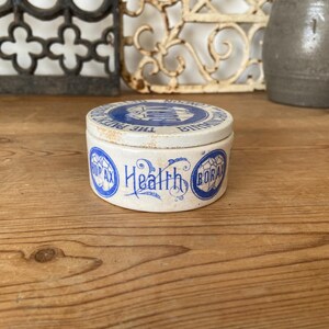 May include: A small, round, antique white ceramic container with a blue lid and blue text. The lid reads "THE PUREST BORAX BIRMINGHAM". The container's side features the words "Health Borax" in blue script.