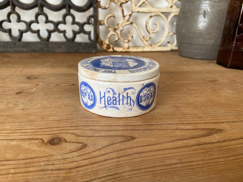 May include: A small, round, antique white ceramic container with a blue lid and blue text. The lid reads "THE PUREST BORAX BIRMINGHAM". The container's side features the words "Health Borax" in blue script.