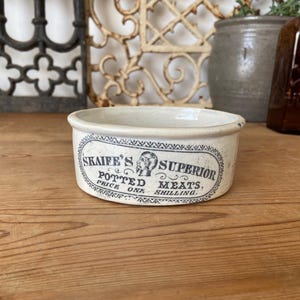 May include: Antique, off-white ceramic Skaife's Superior Potted Meats dish. The oval label features text and a portrait. The dish is on a wooden surface. Other vintage items are in the background.