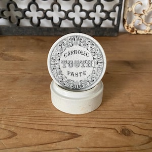 Könnte beinhalten: Ein runder, cremeweißer Keramikbehälter mit einem Deckel, der einen schwarzen dekorativen Rand und den Text "CARBOLIC TOOTH PASTE" in der Mitte aufweist. Der Deckel hat eine sichtbare Rissglasur und der Boden ist ein einfacher Zylinder.