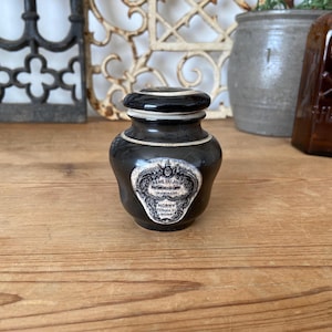 May include: A small, dark brown antique jar with a black lid and a white label. The label has ornate detailing and text, including the words "CREME DU JOUR" and "HORNY". The jar is on a wooden surface.