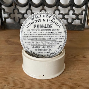 Könnte beinhalten: Ein runder weißer Keramikbehälter mit einem Deckel, der mit schwarzem Text bedruckt ist: WILLETT'S NUTRITIVE & SEDATIVE POMADE, FOR PREVENTING & RESTORING GREY HAIR, ALLAYS ALL IRRITATION OF THE SKIN OF THE HEAD, AND RENDERS THE HAIR SOFT AND GLOSSY.