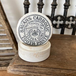 Könnte beinhalten: Ein runder, weißer Keramikbehälter mit einem Deckel, der den schwarzen Aufdruck Savars Carbolic Tooth Paste for Cleaning and Preserving the Teeth & Gums, ein zentrales dreieckiges Logo und eine sichtbare Craquelé-Glasur aufweist.