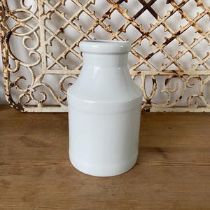 Puede incluir: Una botella de leche de cerámica blanca con un fondo redondeado y una parte superior ligeramente acampanada. La botella está sobre una superficie de madera.