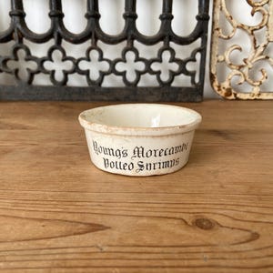 Pode incluir: Um pequeno pote de cerâmica redondo, de cor branco-sujo, com uma borda ligeiramente desgastada. O exterior apresenta um texto preto numa fonte decorativa que diz Young's Morecambe Potted Shrimps. O recipiente tem um interior liso.