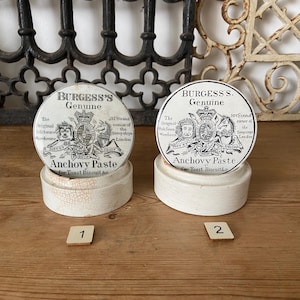 Peut inclure: Deux pots de pâte d'anchois Burgess's Genuine vintage. Chaque pot a une base en céramique blanche et un couvercle rond avec une étiquette en noir et blanc. Les pots sont sur une surface en bois.