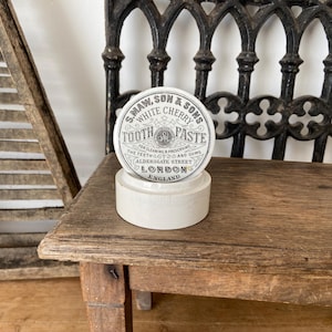Könnte beinhalten: Ein runder weißer Keramikbehälter mit einem Deckel, der mit schwarzem Text bedruckt ist: S. MAW, SON & SONS WHITE CHERRY TOOTH PASTE FOR CLEANING & PRESERVING THE TEETH AND GUMS ALDERSGATE STREET LONDON ENGLAND. Der Behälter hat eine glatte Oberfläche.