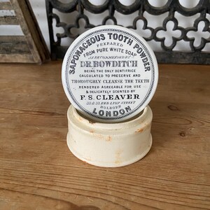 Könnte beinhalten: Ein runder, cremefarbener Keramikbehälter mit einem Deckel, der den schwarzen Aufdruck Saponaceous Tooth Powder, Prepared from Pure White Soap, as recommended by Dr. Bowditch, F. S. Cleaver, 32 & 33 Red Lion Street, Holborn, London trägt.