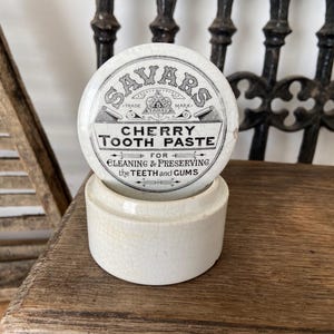 May include: A round, off-white ceramic pot with a matching lid featuring black printed text that reads Savars Cherry Tooth Paste for Cleaning & Preserving the Teeth and Gums. The surface of the ceramic shows a fine, aged crackle glaze finish.