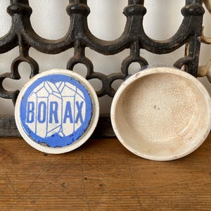 May include: An open, antique ceramic container with a lid. The lid is white with a blue border and the word "BORAX" in blue. The container is off-white with a cracked glaze.