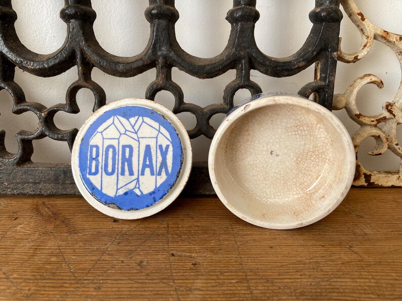 May include: An open, antique ceramic container with a lid. The lid is white with a blue border and the word "BORAX" in blue. The container is off-white with a cracked glaze.