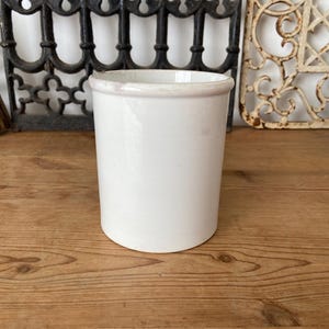 Puede incluir: Una vasija cilíndrica de cerámica blanca con un acabado liso y brillante y un borde ligeramente engrosado. El recipiente presenta un diseño sencillo y sin adornos con un color blanco uniforme, mostrando una estructura limpia y funcional.