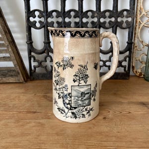 Pode incluir: Uma jarra de cerâmica cor de creme com um padrão de transferência preto de ramos florais e uma cena paisagística central que retrata um castelo junto à água. A borda é decorada com um motivo de folhas e a pega tem uma textura.