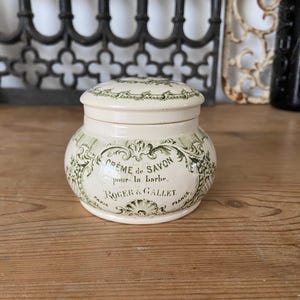 May include: A cream-coloured ceramic jar with a matching lid, featuring green decorative scrollwork and text that reads CRÈME de SAVON pour la barbe ROGER & GALLET PARIS FRANCE. The container has a rounded body and a smooth, glossy finish.