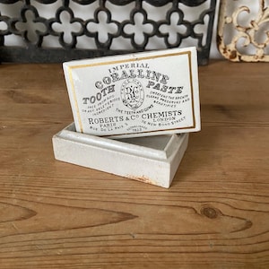 Könnte beinhalten: Eine rechteckige weiße Keramikdose mit einem Deckel, der schwarzen Text mit der Aufschrift Imperial Coralline Tooth Paste, Roberts & Co Chemists, London, Paris und 76 New Bond Street sowie einen goldenen Rand und ein Logo trägt.