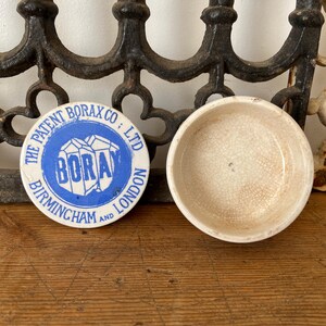 May include: An antique ceramic bowl and a round lid with blue text that reads "The Patent Borax Co; Ltd, Borax, Birmingham and London". The bowl is off-white with a cracked glaze. The items are on a wooden surface.