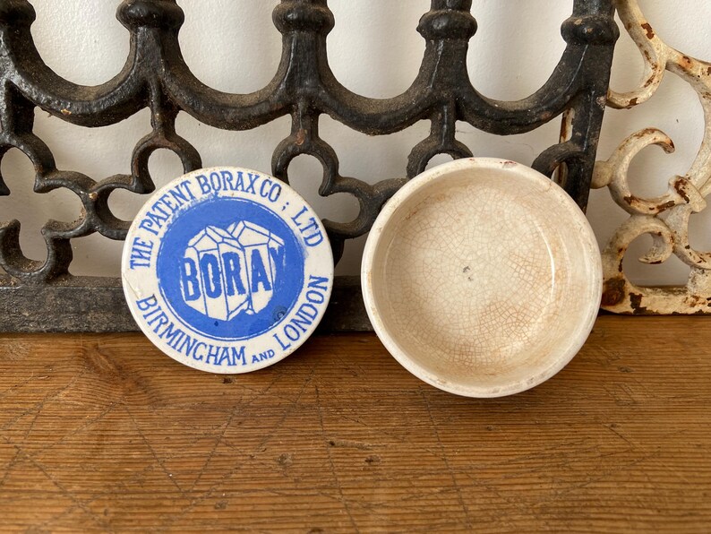 May include: An antique ceramic bowl and a round lid with blue text that reads "The Patent Borax Co; Ltd, Borax, Birmingham and London". The bowl is off-white with a cracked glaze. The items are on a wooden surface.