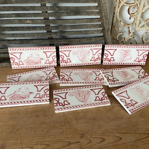 Può includere: Otto piastrelle in ceramica rossa e bianca con un motivo ripetuto di testa di animale stilizzata. Le piastrelle presentano un design di mucca, pecora e toro.