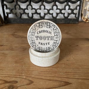 Könnte beinhalten: Ein rundes, cremeweißes Keramikgefäß mit passendem Deckel, verziert mit einem dekorativen schwarzen Schnörkelrand und dem mittig aufgedruckten Text "CARBOLIC TOOTH PASTE". Die Keramikoberfläche weist ein feines, sichtbares Netz aus Glasur-Rissen auf.