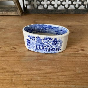 以下が含まれることがあります： 青と白のウィローパターンが施された楕円形の陶器製ディッシュ。外側には、パゴダ、木々、風景要素の詳細な転写プリントが施されています。内側は白で、上部の縁に沿って青い模様のボーダーがあります。