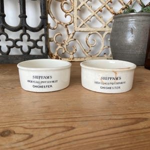 May include: Two antique, off-white ceramic bowls with the text "SHIPPAM'S HIGH CLASS POTTED MEAT CHICHESTER" in black lettering. The bowls are round with a slightly flared rim. The background includes decorative metalwork and a brown bottle.