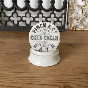 Könnte beinhalten: Ein weißer Keramikbehälter mit Deckel, der mit dem schwarzen Aufdruck PINCH & CO. COLD CREAM BATH und dekorativen Schnörkeln versehen ist. Der Behälter verfügt über einen runden Boden und einen passenden Deckel in schlichtem Design.