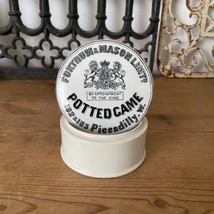Può includere: Un barattolo in ceramica color crema di forma rotonda con un coperchio decorato da testo nero e uno stemma reale. Il coperchio riporta le scritte FORTNUM & MASON LIMT, POTTED GAME, BY APPOINTMENT TO THE KING e 182 & 183 PICCADILLY. W. Base cilindrica.