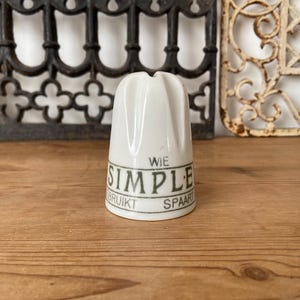 以下が含まれることがあります： 上部が溝付きの白いセラミック製シンブルで、緑色の文字で「WIE SIMPLE GEBRUIKT SPAART」と印刷されています。シンブルは円筒形で、本体はわずかに先細りになっており、上部中央には針を通すための小さな穴が開いています。