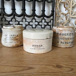May include: Three cylindrical ceramic pots with lids, featuring a cream-coloured crackle glaze finish. Each container displays black printed text, including names like CH CARLE, ENDLE CHEMIST, and P. TRUEFITT, along with various location details.
