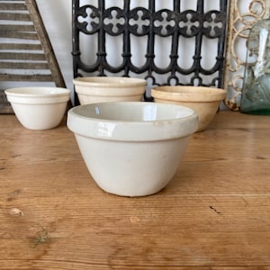May include: A collection of three off-white ceramic mixing bowls with a smooth finish and a distinct rimmed edge. The bowls are arranged in a cluster, featuring a rounded base and a wide opening, showcasing a simple, functional design for kitchen use.