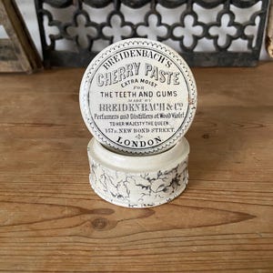 Könnte beinhalten: Ein runder, cremeweißer Keramiktopf mit einem Deckel, der den schwarzen Text trägt: BREIDENBACH'S CHERRY PASTE EXTRA MOIST FOR THE TEETH AND GUMS MADE BY BREIDENBACH & CO PERFUMERS AND DISTILLERS OF WOOD VIOLET TO HER MAJESTY THE QUEEN 157B NEW BOND STREET LONDON.