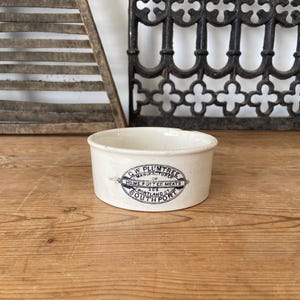 Puede incluir: Un pequeño recipiente de cerámica de color blanco roto con una etiqueta impresa en negro en la parte frontal. El texto dice C.W. PLUMTREE MANUFACTURER OF HOME POTTED MEATS 206 PORTLAND ST SOUTHPORT. El cuenco tiene un borde liso y base plana.