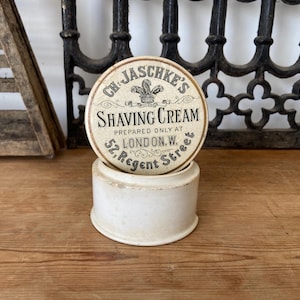 May include: A round, off-white ceramic shaving cream container with a lid. The lid features black text that reads: CH. JASCHKE'S SHAVING CREAM PREPARED ONLY AT LONDON. W. 52, REGENT STREET, along with a small graphic of a plume emblem.