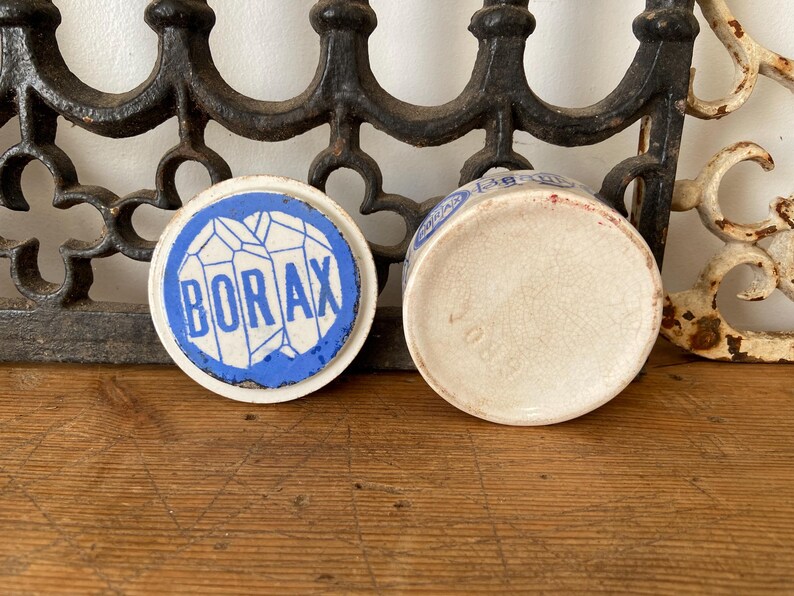 May include: An antique ceramic Borax container with a blue and white design. The lid features the word "BORAX" in a bold font, surrounded by a blue border. The container is off-white with a cracked texture.