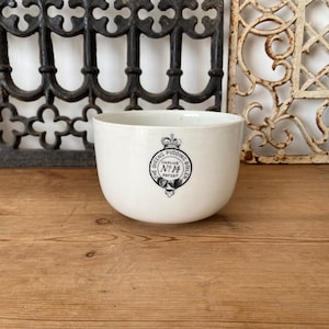 Puede incluir: Un cuenco de cerámica blanca con un emblema circular negro en la parte frontal. El emblema muestra un icono de corona sobre el texto THE QUEENS PUDDING BOILER, con CHALUG NO 14 PATENT impreso en el centro. El cuenco tiene un exterior liso.
