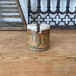 Op de afbeelding: Een cilindrische keramische Winsor & Newton houder met een wit deksel en een metalen clip. De behuizing heeft een beige etiket met een groen en goudkleurige illustratie van een gevleugelde leeuw, zwarte tekst en de merknaam Winsor & Newton Ltd.