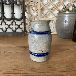 May include: A small, antique stoneware jar with a light blue and grey speckled finish. The jar has two dark blue bands and embossed text. The jar is on a wooden surface.